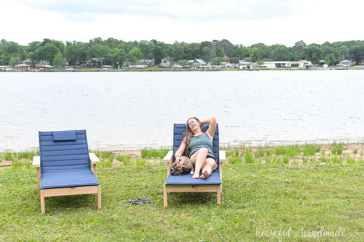 Girl and dog relaxing on a wooden chaise lounge chair with navy cushion in front of a lake. 