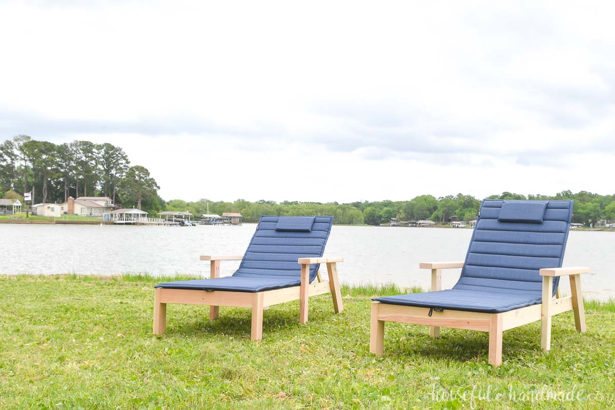 Two pool lounge chairs with thin cushions on the lawn by a lake. 