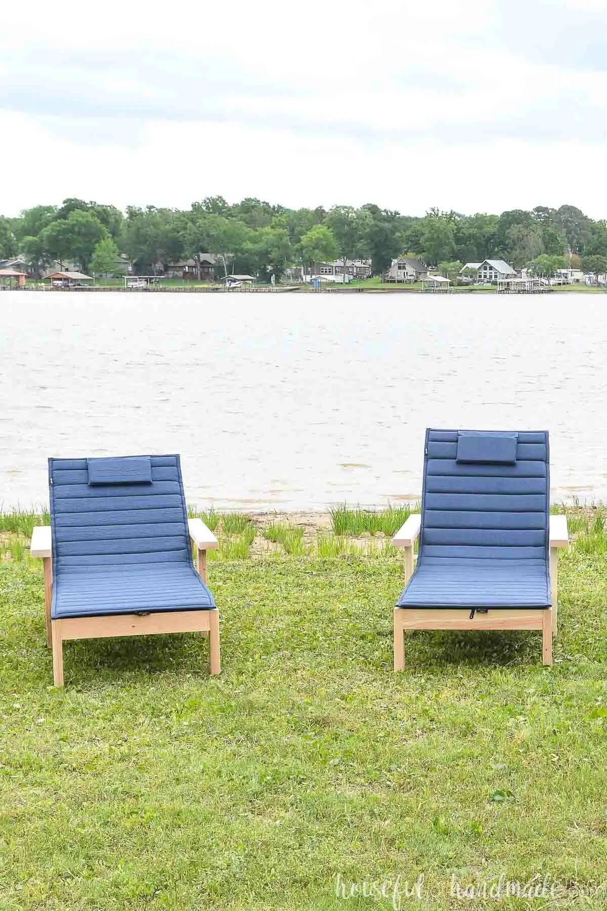 Two lounge chairs with thin, roll-up cushions, sitting in the grass by a lake. 