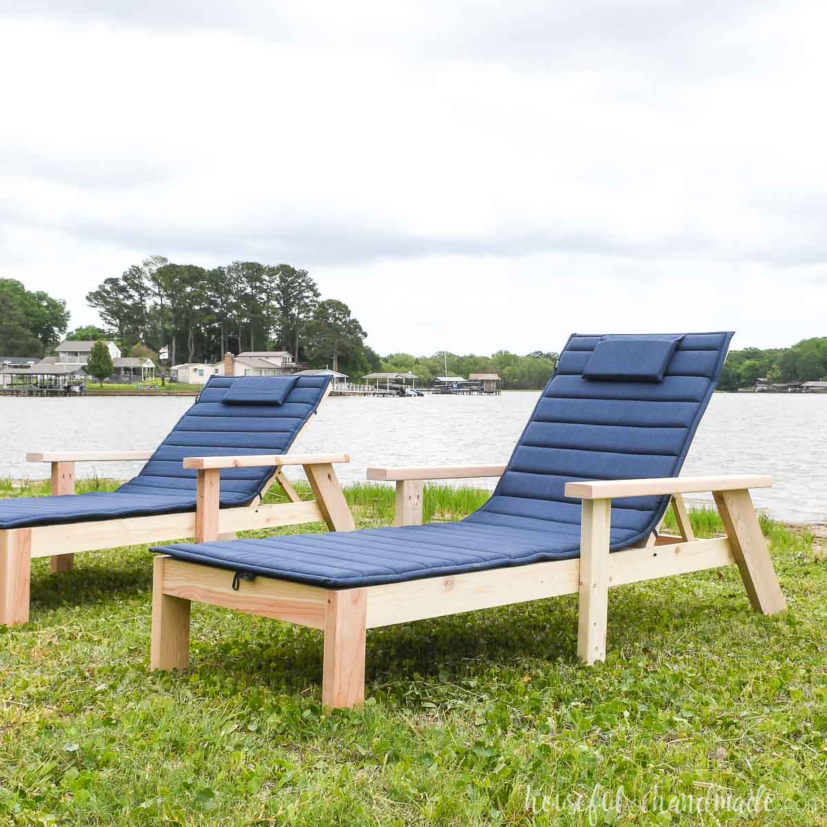 Two wood pool chaise lounge chairs on the grass next to a lake.