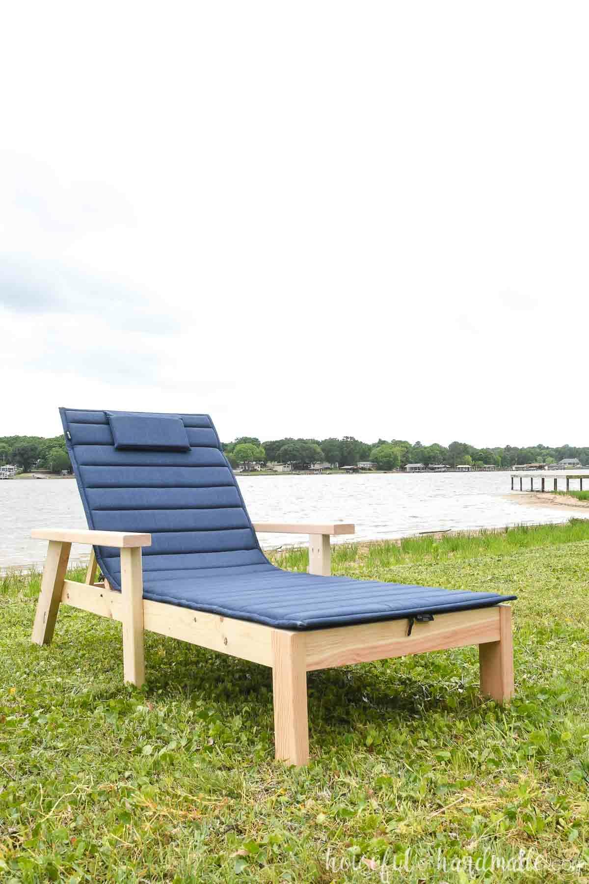 Wood chaise lounge chair with navy cushion on a lawn in front of a lake.