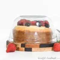 White cake with berries on wood cake plate made from scrap wood with glass dome.