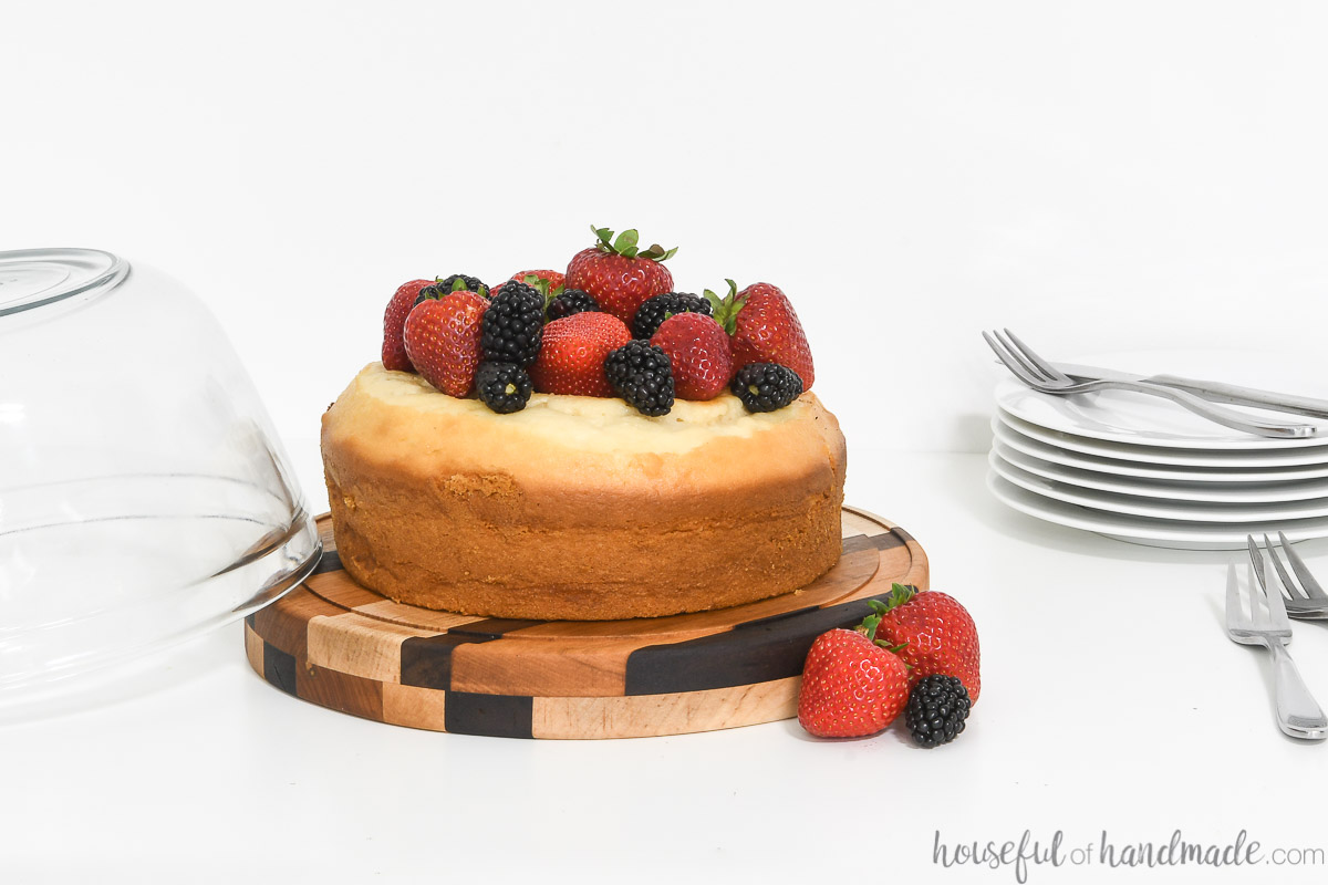 White cake with strawberries and blackberries on a wood cake plate made from scraps of hardwoods with a stack of serving plates in the back. 
