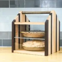 Wood pie carrier with room for 2 pies, dowel handle, and acrylic front on a counter with 2 pies in it.