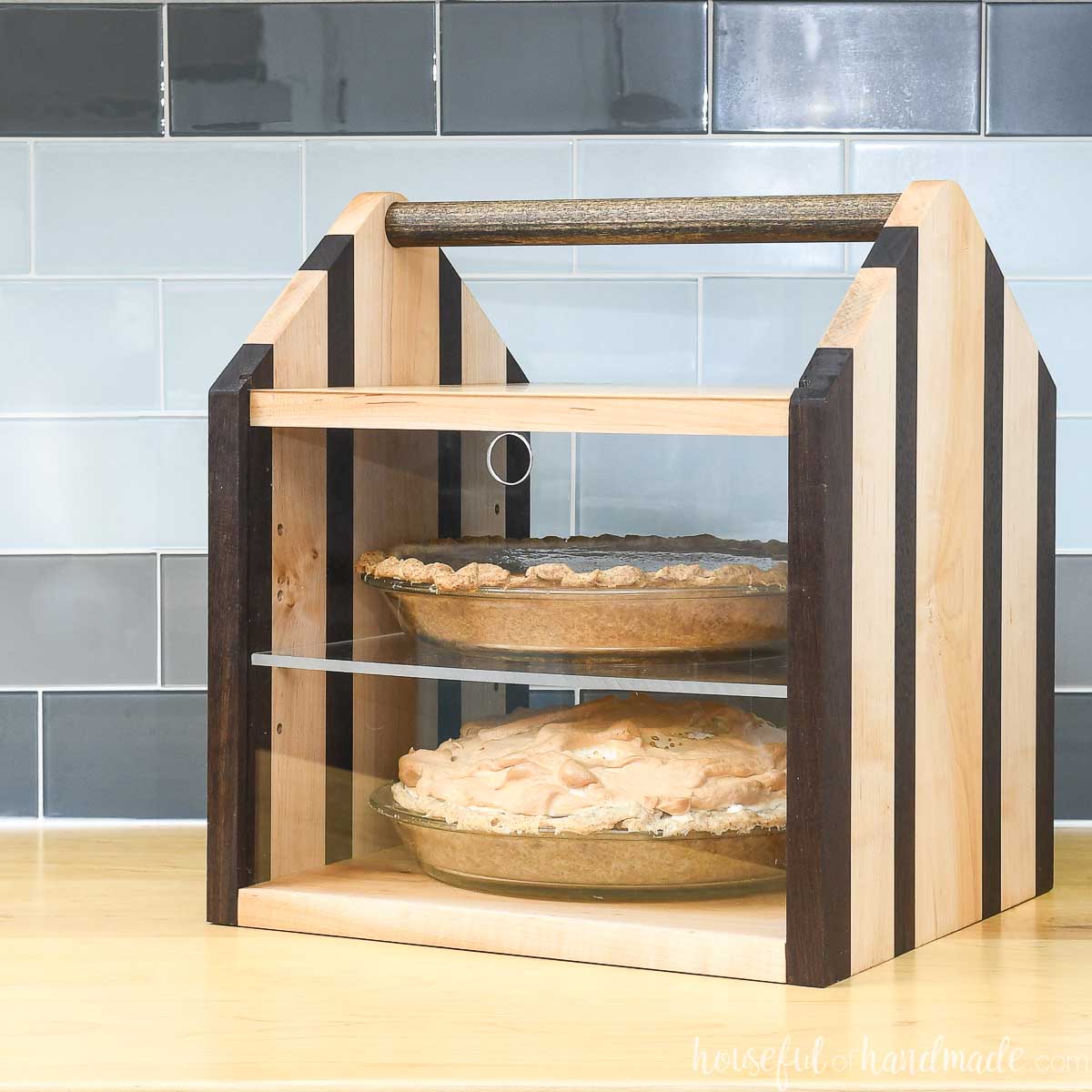 Wood pie carrier with room for 2 pies, dowel handle, and acrylic front on a counter with 2 pies in it.