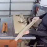 Tapered leg held up in front of a table saw with a DIY tapered leg jig sitting on it.