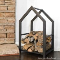 Black firewood holder with a modern house shape built out of wood next to a fireplace.
