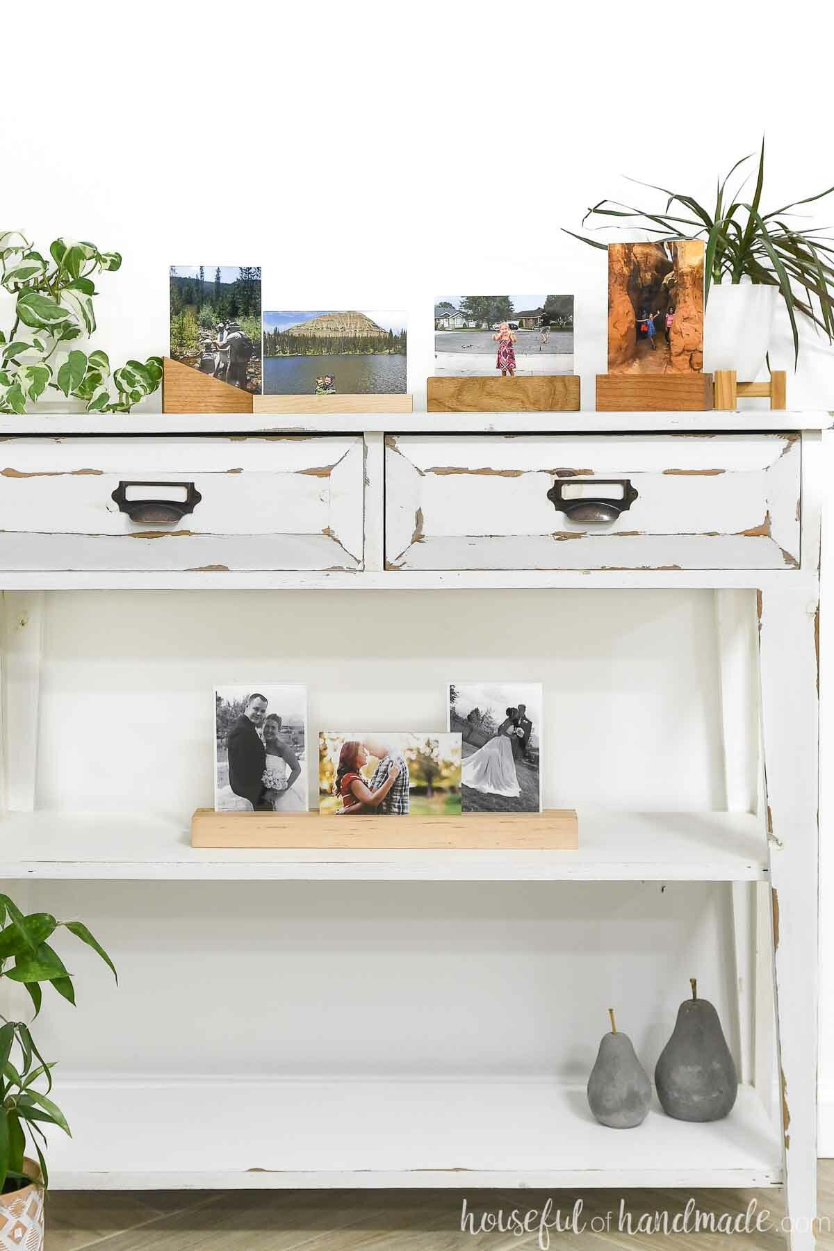 White console table filled with pictures held up by wood block frames with plants in the back. 