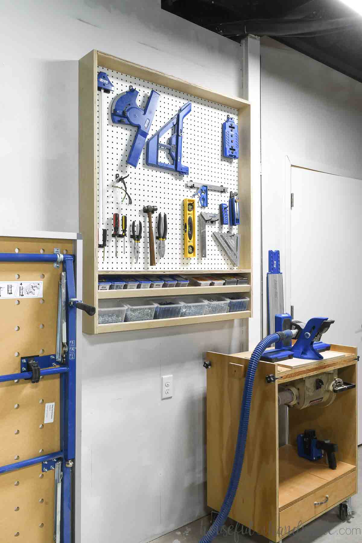 Pegboard storage cabinet hanging on a wall in a garage with tools on it and larger tools next to it.