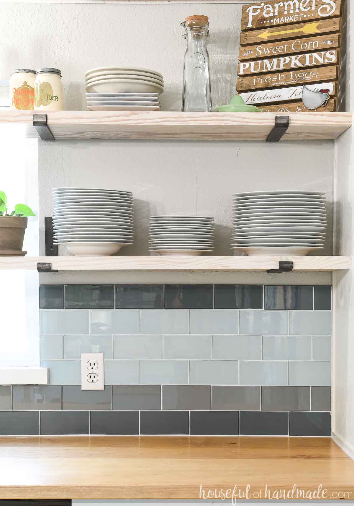 Corner of a kitchen with open shelves holding platesand glass tile backsplash.