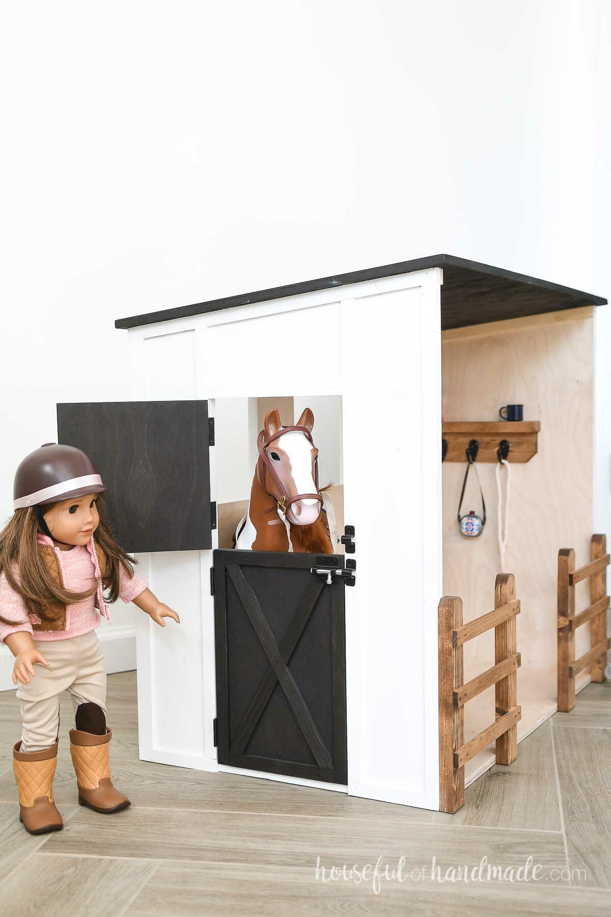 Side of the horse barn with a black stained dutch door half opened and a toy horse sticking it's head out with an 18" doll on the outside. 