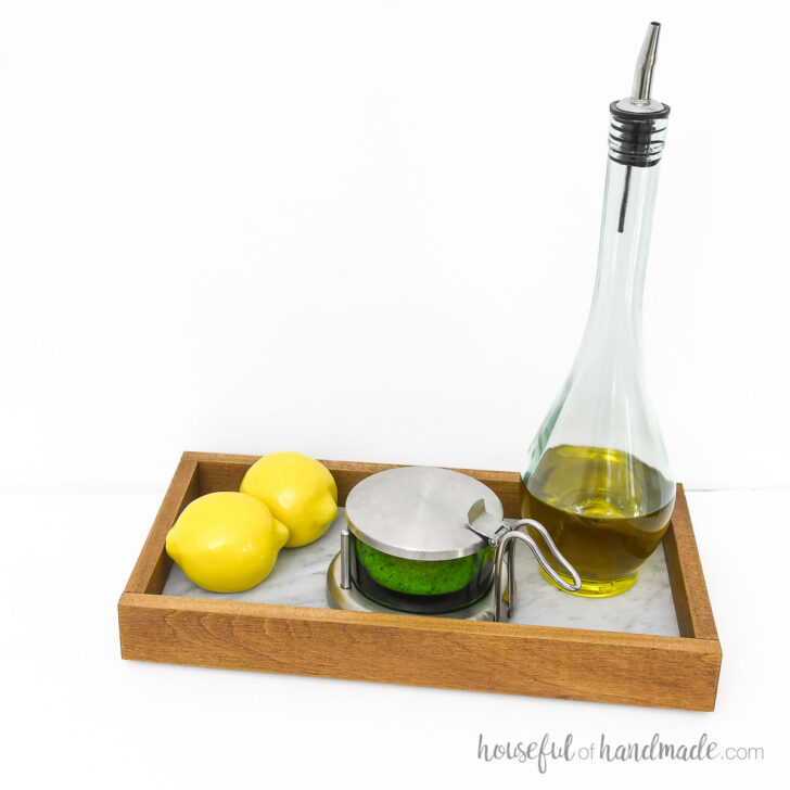 Organizing tray with oil bottle, salt cellar and lemons in it on a white surface.