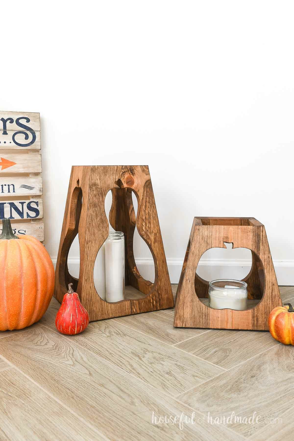 Two tapered lanterns with pumpkin shapes surrounded by pumpkins next to a wood sign.