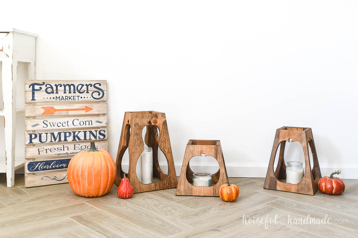 Three different sized wooden fall lanterns in a display with a fall sign and pumpkins.