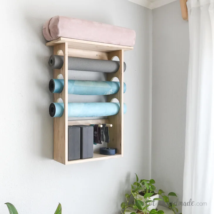 DIY yoga mat holder mounted on a wall with blue mats and a pink bolster stored in it.