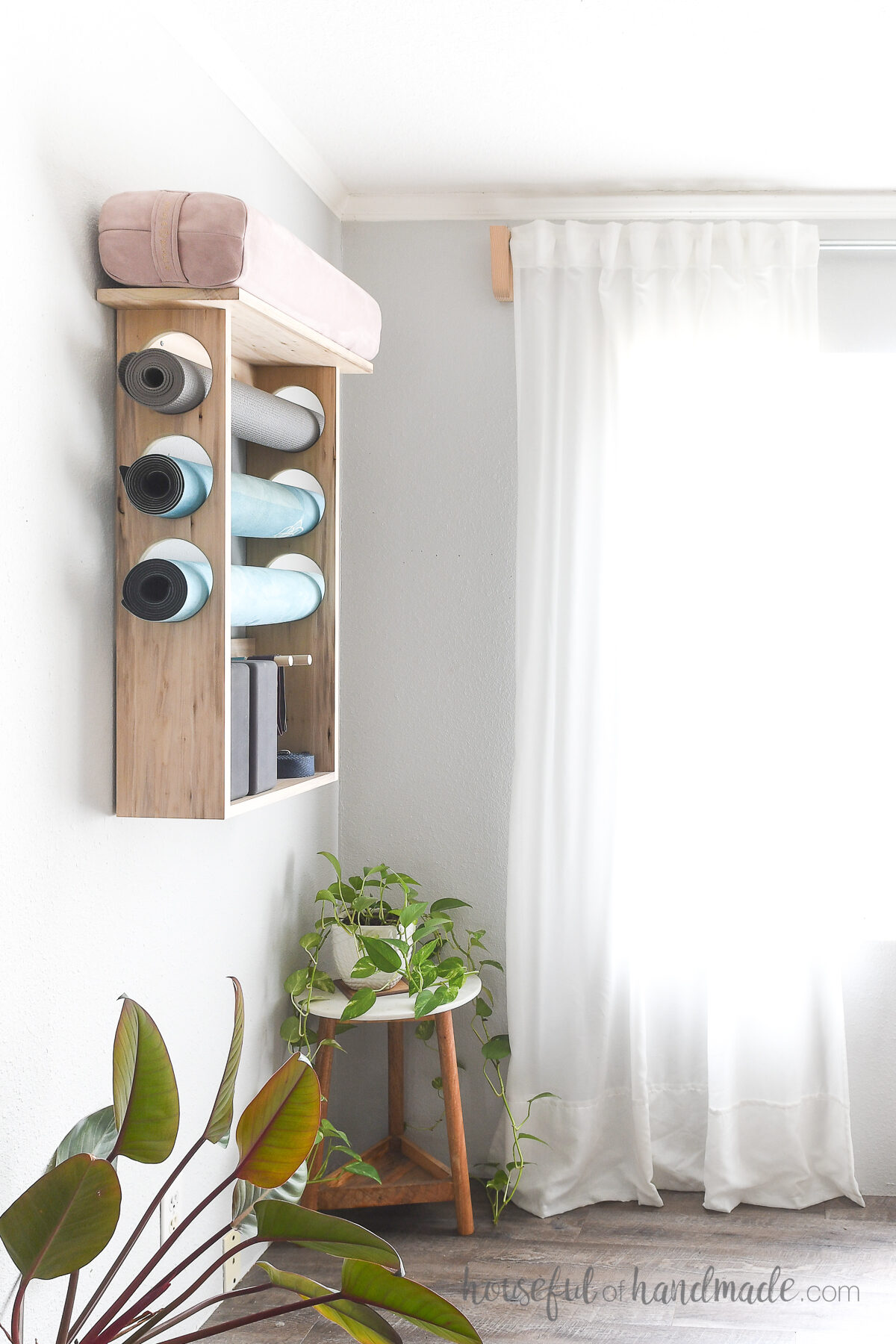 Wood DIY yoga mat holder on the wall with 3 yoga mats inside and plants on the floor by it. 