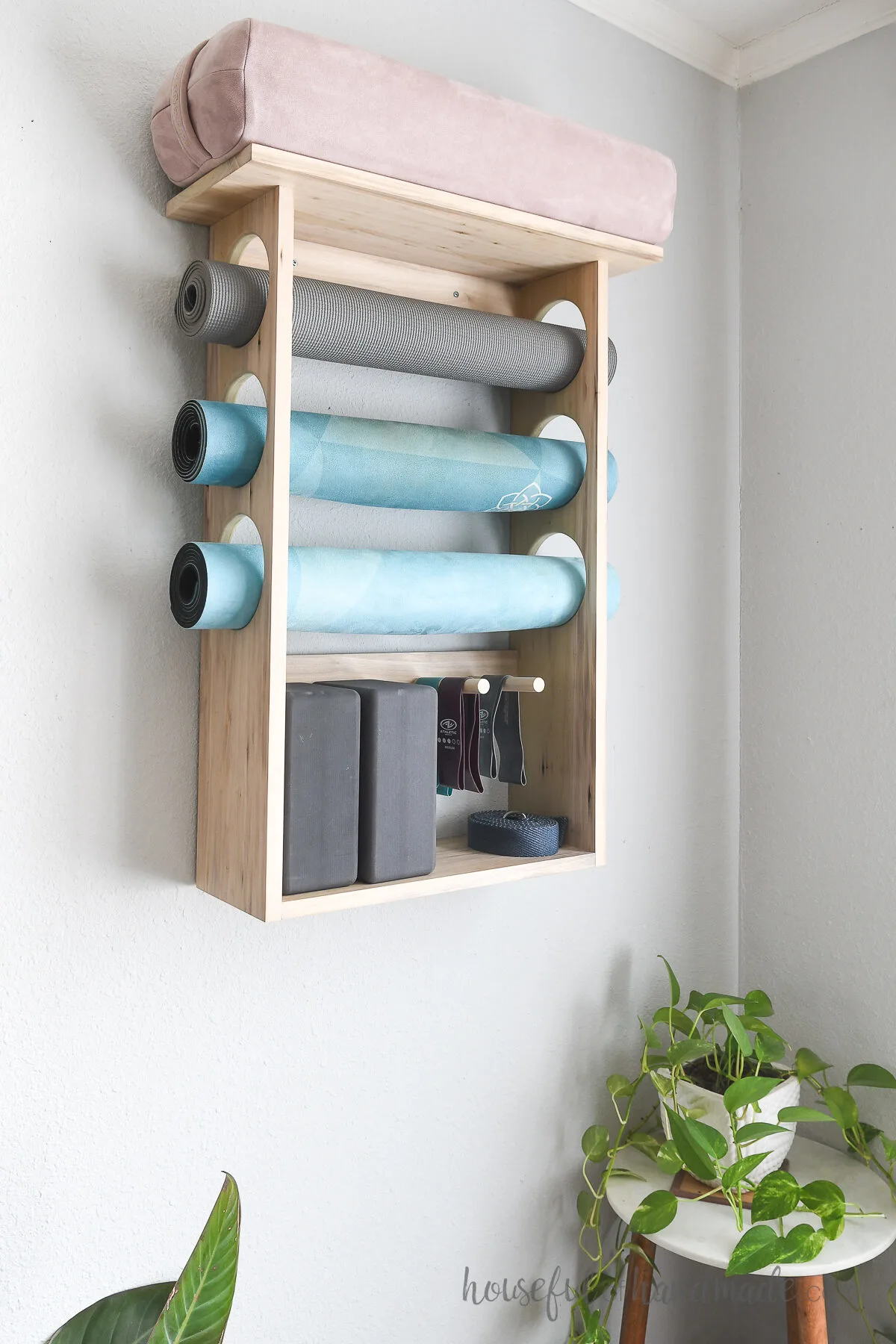 Three yoga mats in a wall mounted holder with a bolster on the top and blocks below. 
