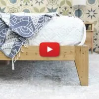 DIY wood bed frame with mattress with white blanket with play button in the center.
