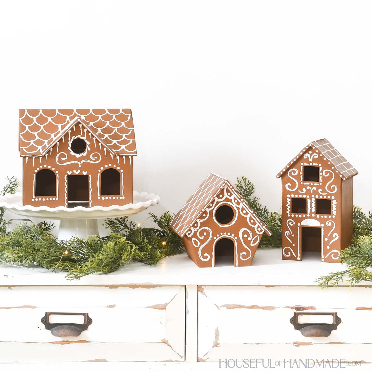 Three DIY wood gingerbread houses on a table with garland around them.